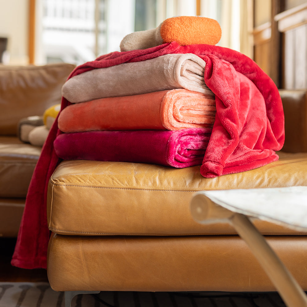 A stack of 4 bright pink, orange, and red VelvetLoft Throws sitting on the edge of an orange leather sofa.