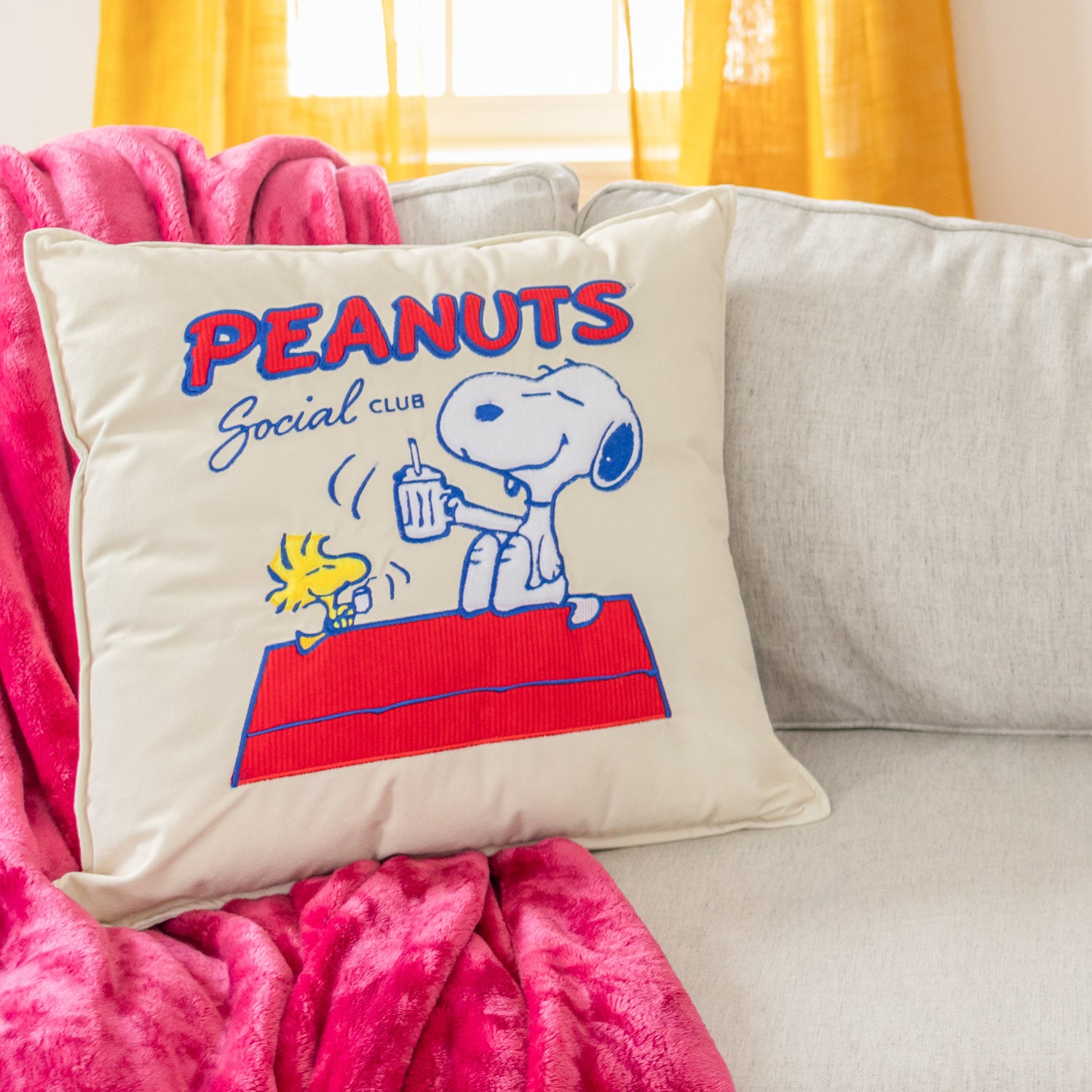 Peanuts-themed pillow with Snoopy and Woodstock on a couch with pink blanket and yellow curtains.