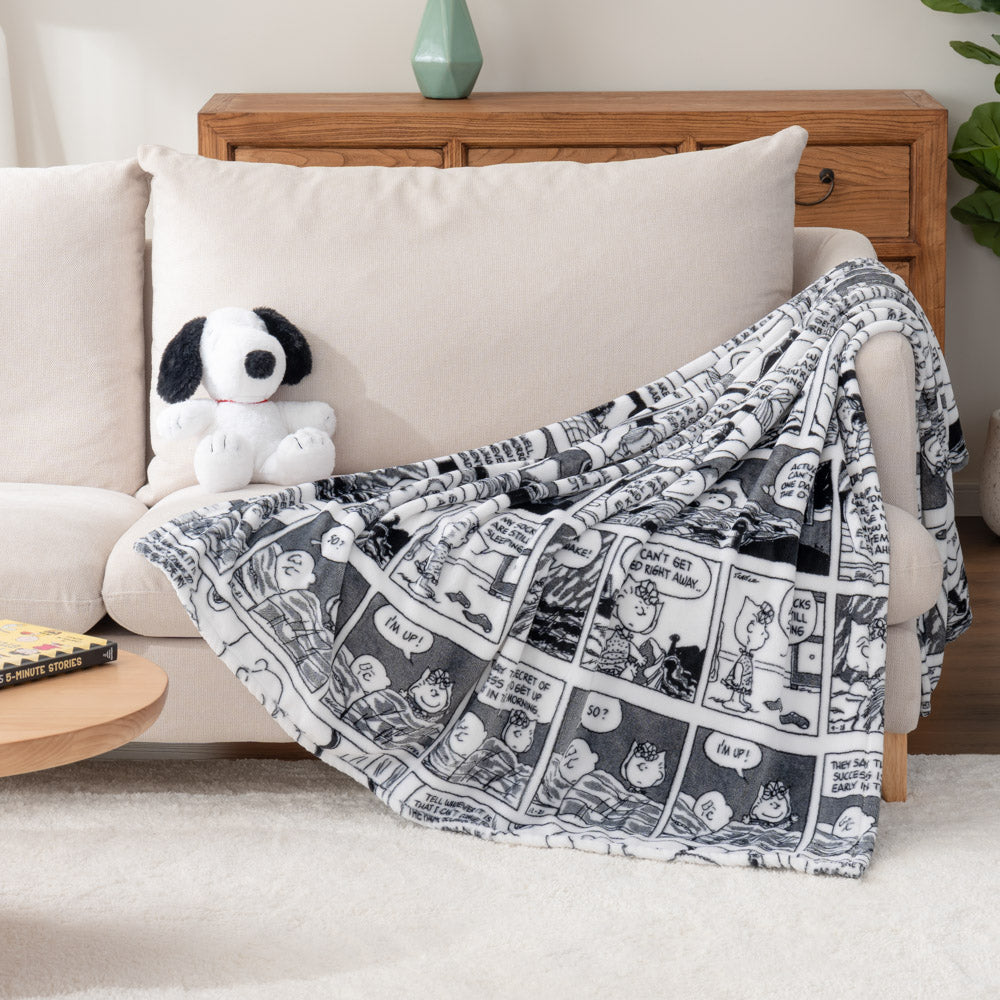 A white throw blanket with Peanuts comic strip illustrations draped over an armchair with a snoopy plush toy sitting beside it.