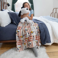 Person holding a Snoopy plush toy on a bed with a Snoopy-themed blanket