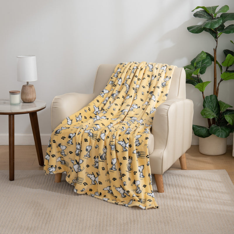 Yellow peanuts poses and paws throw draped neatly over a beige leather chair in a room next to a table and plant.