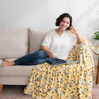 A woman sitting on a sofa as she leans on a yellow peanuts poses and paws throw draped over a beige sofa.