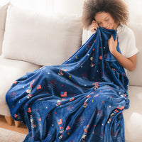 A cobalt blue Peanuts flag parade throw draped over a womans legs as she snuggles with it while smiling.