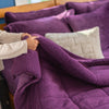 A person pulling a purple quilted comforter over themselves on a bed, creating a cozy scene.