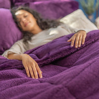 A person lounging on a bed covered with a purple quilted comforter, smiling and looking relaxed.