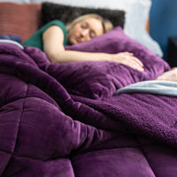 A person lying on a bed covered with a purple quilted comforter, holding a pink pillow and smiling.