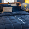 A navy quilted comforter on a bed with navy pillows and beige cushions in a warmly lit bedroom.