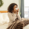 Woman sitting on a bed holding a mug, wrapped in a brown blanket in a cozy room.