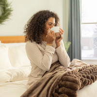 Woman sitting on a bed holding a mug, wrapped in a brown blanket in a cozy room.