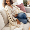 Woman sitting on a couch wrapped in a chunky knit blanket, wearing a white sweater and blue jeans.
