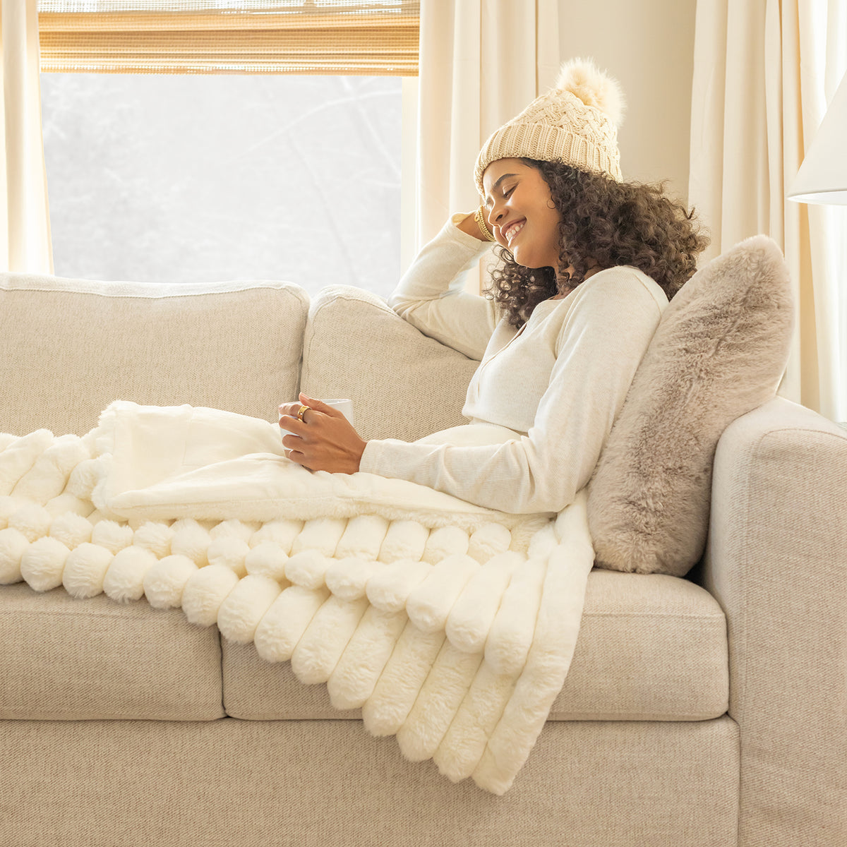 Woman sitting on a couch with a white textured blanket and beige hat, surrounded by soft lighting.