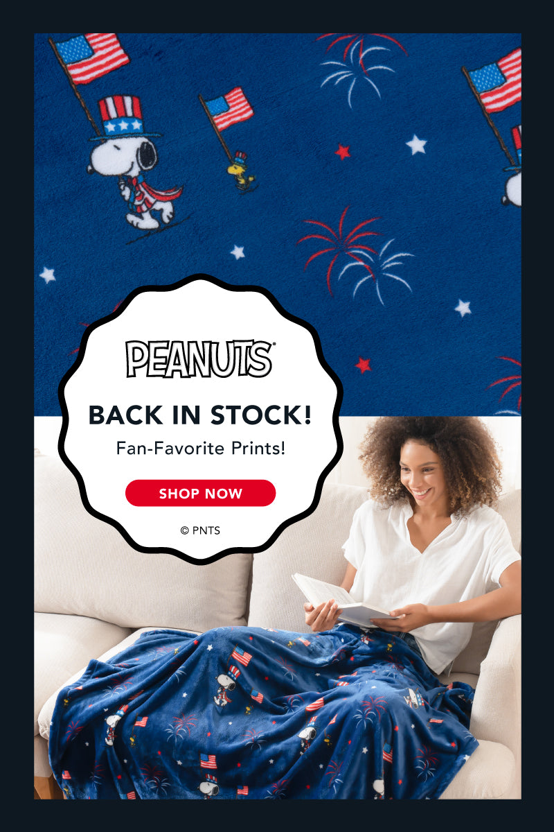 Peanuts® - BACK INS TOCK! Fan-Favorite Prints! Shop Now. Image of a woman enjoying a cup of tea while using her Snoopy and Woodstock Americana print throw.