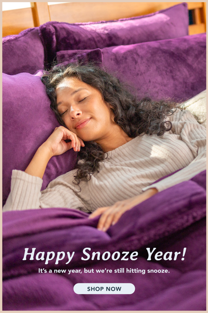 Relaxing bedroom scene with a woman peacefully sleeping under a luxurious deep purple blanket. The bedding setup features matching purple pillows and a plush cream accent pillow for added texture. The text reads, 'Happy Snooze Year! It’s a new year, but we’re still hitting snooze,' with a call-to-action button saying 'Shop Now,' inviting viewers to indulge in cozy comfort."