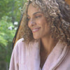 A video of a woman smiling outdoors, wearing a pink VelvetLoft Robe while she stands next to a sauna.