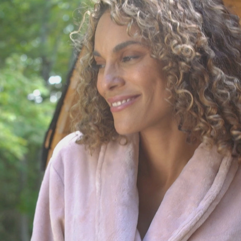 A video of a woman smiling outdoors, wearing a pink VelvetLoft Robe while she stands next to a sauna.